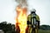 A firefighter wearing protective gear stands facing a large, intense fire with flames and smoke billowing upwards. The firefighter's back is towards the camera, and they are equipped with an oxygen tank. The surrounding area includes trees and a small building in the background.