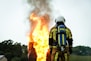 A firefighter wearing protective gear stands facing a large, intense fire with flames and smoke billowing upwards. The firefighter's back is towards the camera, and they are equipped with an oxygen tank. The surrounding area includes trees and a small building in the background.