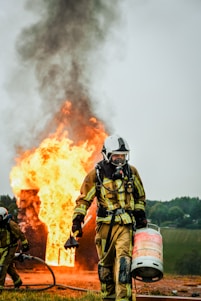 A firefighter in full protective gear carries a propane tank while walking away from a large fire. Flames and thick black smoke rise from a burning structure in the background. Another firefighter is partially visible holding a hose near the fire.