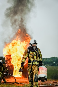 A firefighter in full protective gear carries a propane tank while walking away from a large fire. Flames and thick black smoke rise from a burning structure in the background. Another firefighter is partially visible holding a hose near the fire.