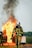A firefighter in full protective gear carries a propane tank while walking away from a large fire. Flames and thick black smoke rise from a burning structure in the background. Another firefighter is partially visible holding a hose near the fire.