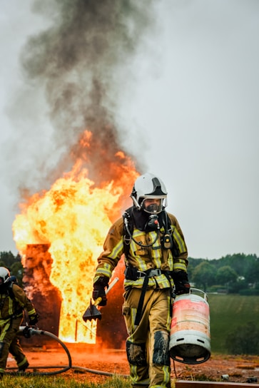A firefighter in full protective gear carries a propane tank while walking away from a large fire. Flames and thick black smoke rise from a burning structure in the background. Another firefighter is partially visible holding a hose near the fire.