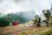Firefighters in full gear, including helmets and oxygen tanks, are positioned on the ground near hoses, facing a scene with smoke rising in the background. The setting is outdoors, with a mix of grassy and dirt terrain, and a forested area providing a backdrop. A red and gray armchair sits in stark contrast amidst the scenery. A yellow vehicle is visible in the background.