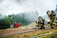 Firefighters in full gear, including helmets and oxygen tanks, are positioned on the ground near hoses, facing a scene with smoke rising in the background. The setting is outdoors, with a mix of grassy and dirt terrain, and a forested area providing a backdrop. A red and gray armchair sits in stark contrast amidst the scenery. A yellow vehicle is visible in the background.