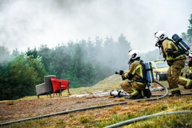 Firefighters in full gear, including helmets and oxygen tanks, are positioned on the ground near hoses, facing a scene with smoke rising in the background. The setting is outdoors, with a mix of grassy and dirt terrain, and a forested area providing a backdrop. A red and gray armchair sits in stark contrast amidst the scenery. A yellow vehicle is visible in the background.