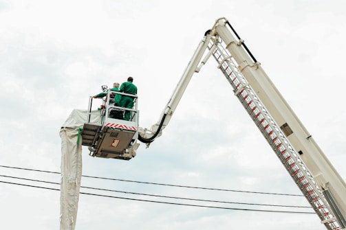 Two individuals wearing green work suits are on an elevated platform attached to a large crane. The crane is extended high above, with support cables visible in the background.