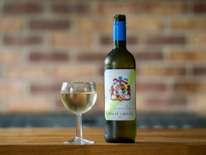 A bottle of Pinot Grigio wine with a colorful label stands next to a filled wine glass on a wooden surface. The background features a blurred brick wall, giving a warm and rustic ambiance.