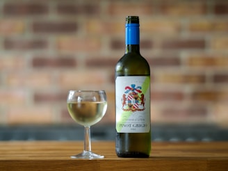 A bottle of Pinot Grigio wine with a colorful label stands next to a filled wine glass on a wooden surface. The background features a blurred brick wall, giving a warm and rustic ambiance.