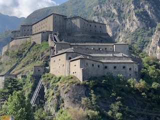 A large, historical fortress is built into the side of a rocky mountain, surrounded by lush greenery. The structure is multi-leveled with numerous windows and stone walls. A steep staircase or ramp leads up the hillside towards the fortress.