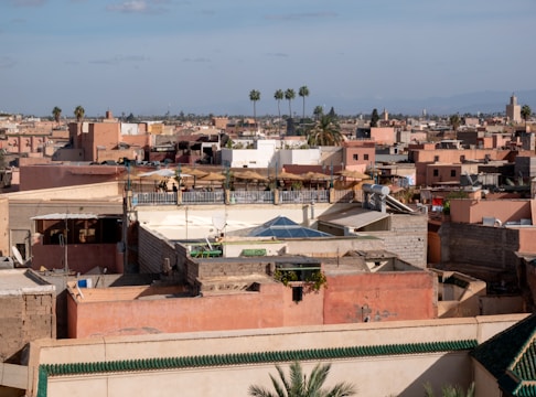 A cityscape featuring a dense cluster of flat-roofed buildings in earthy tones. Several palm trees are visible in the background under a clear sky. A rooftop terrace with umbrellas is noticeable, and some satellite dishes are scattered across the rooftops.