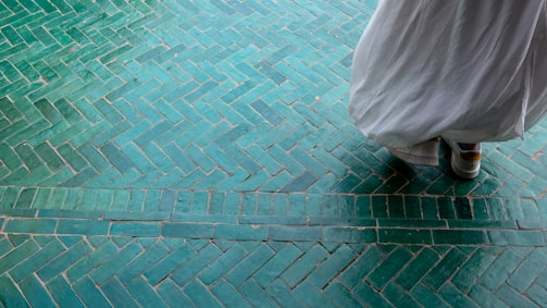 A person wearing a flowing white garment stands on a patterned green tiled floor. The tiles are arranged in a herringbone pattern and create an intricate design with varying shades of green.