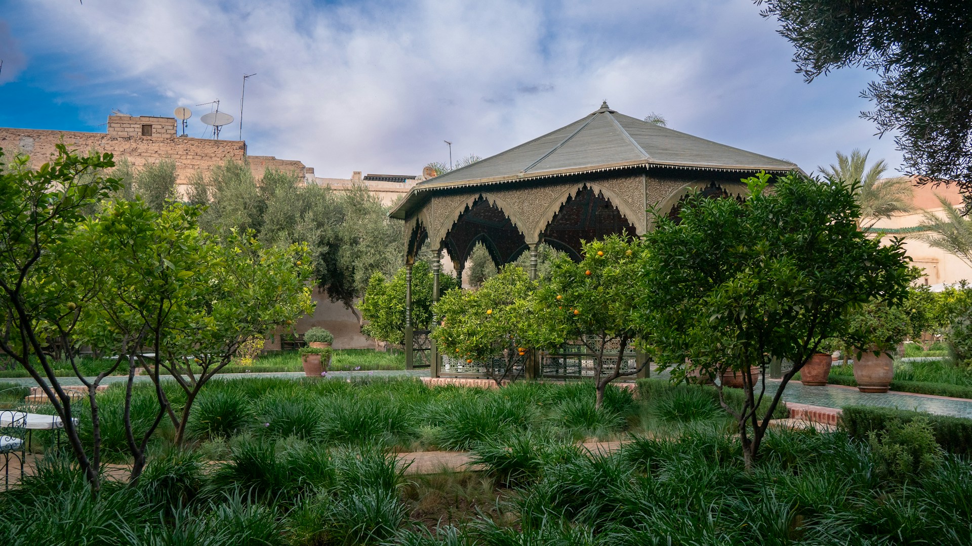 An ornate gazebo is surrounded by lush greenery and citrus trees in a well-maintained garden. Clay pots are scattered throughout the garden, adding to the rustic charm. The sky is partially cloudy, and there are old building walls in the background with satellite dishes on top.