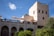 An elegant stone building with multiple levels and arched windows, featuring a terrace area with white umbrellas. The architecture reflects traditional Moroccan design elements. There are people standing on the terrace, and the structure is surrounded by lush greenery. The sky is clear with a few clouds, providing a bright backdrop.