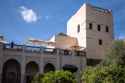 An elegant stone building with multiple levels and arched windows, featuring a terrace area with white umbrellas. The architecture reflects traditional Moroccan design elements. There are people standing on the terrace, and the structure is surrounded by lush greenery. The sky is clear with a few clouds, providing a bright backdrop.