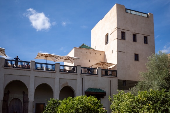 An elegant stone building with multiple levels and arched windows, featuring a terrace area with white umbrellas. The architecture reflects traditional Moroccan design elements. There are people standing on the terrace, and the structure is surrounded by lush greenery. The sky is clear with a few clouds, providing a bright backdrop.