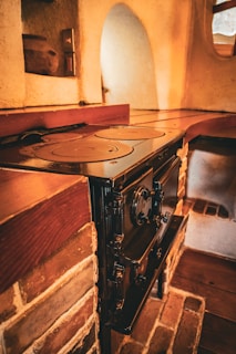 A rustic kitchen scene featuring an old-fashioned cast iron stove set against a brick wall. The stove has multiple circular heating plates on top and a heavy, ornate door. The surrounding area shows the warm and textured surfaces of the bricks and plaster, with a wooden mantel shelf above. Light softly illuminates the space, giving it a cozy and vintage feel.