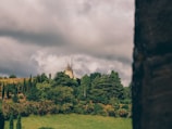 A rustic windmill is situated on a lush, green hill surrounded by dense trees and tall, slender bushes. The sky is overcast with thick, gray clouds, contributing to a moody and serene atmosphere.