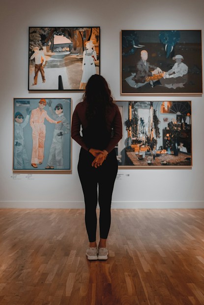 A person stands in an art gallery, facing a wall with four paintings displayed. The atmosphere is calm, with warm lighting highlighting each artwork. The floor is wooden, adding an earthy tone to the scene.