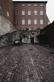 A parked car in a cobblestone courtyard surrounded by stone and brick walls. The building in the background is multi-story with multiple windows. Vines are growing on parts of the walls, adding a touch of greenery.