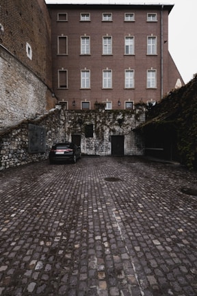 A parked car in a cobblestone courtyard surrounded by stone and brick walls. The building in the background is multi-story with multiple windows. Vines are growing on parts of the walls, adding a touch of greenery.