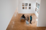 A modern art gallery features various framed artworks displayed along white walls. Two individuals are viewing the art, one standing and the other seated. A third person is blurred, suggesting movement. The floors are made of polished wood, and the atmosphere is serene and contemplative.
