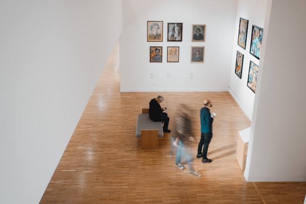 A modern art gallery features various framed artworks displayed along white walls. Two individuals are viewing the art, one standing and the other seated. A third person is blurred, suggesting movement. The floors are made of polished wood, and the atmosphere is serene and contemplative.