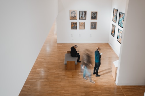 A modern art gallery features various framed artworks displayed along white walls. Two individuals are viewing the art, one standing and the other seated. A third person is blurred, suggesting movement. The floors are made of polished wood, and the atmosphere is serene and contemplative.