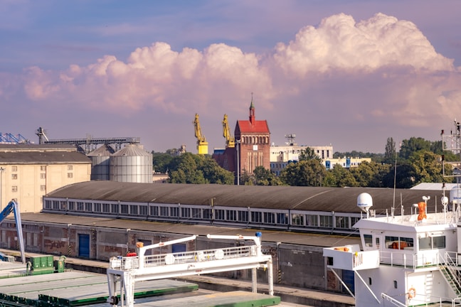 An industrial area features several large buildings, including a long warehouse with a row of windows. Two large yellow cranes are positioned near a red brick building with a pointed rooftop and a tower. In the foreground, a white maritime vessel is docked, showcasing parts of its deck and equipment. Trees and other industrial structures can be seen in the background, and fluffy clouds float gently in a blue sky.