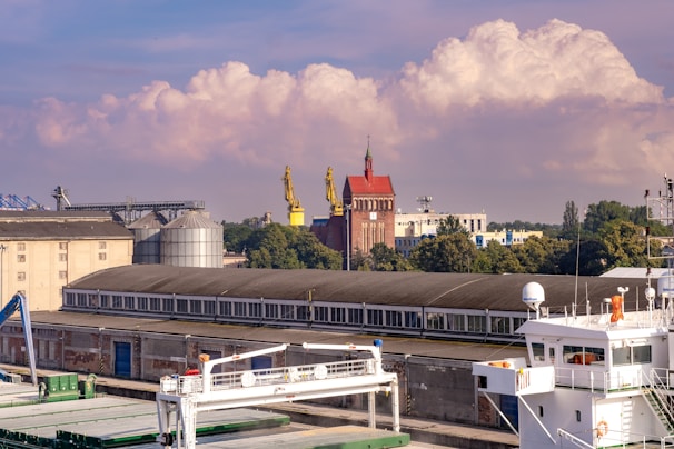 An industrial area features several large buildings, including a long warehouse with a row of windows. Two large yellow cranes are positioned near a red brick building with a pointed rooftop and a tower. In the foreground, a white maritime vessel is docked, showcasing parts of its deck and equipment. Trees and other industrial structures can be seen in the background, and fluffy clouds float gently in a blue sky.