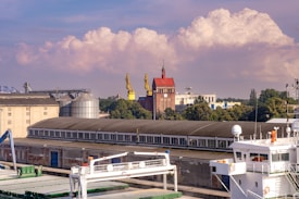 An industrial area features several large buildings, including a long warehouse with a row of windows. Two large yellow cranes are positioned near a red brick building with a pointed rooftop and a tower. In the foreground, a white maritime vessel is docked, showcasing parts of its deck and equipment. Trees and other industrial structures can be seen in the background, and fluffy clouds float gently in a blue sky.
