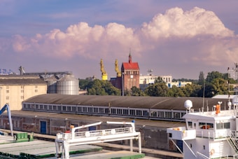 An industrial area features several large buildings, including a long warehouse with a row of windows. Two large yellow cranes are positioned near a red brick building with a pointed rooftop and a tower. In the foreground, a white maritime vessel is docked, showcasing parts of its deck and equipment. Trees and other industrial structures can be seen in the background, and fluffy clouds float gently in a blue sky.