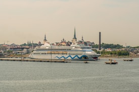 A large cruise ship is docked at a port with a cityscape in the background. The ship features a distinctive design with blue waves and eye motifs on its hull. The city in the background is characterized by several historic buildings with steeples, suggesting a European architectural style. A tall, narrow smokestack is visible, indicating an industrial area nearby. The scene conveys a sense of travel and exploration.