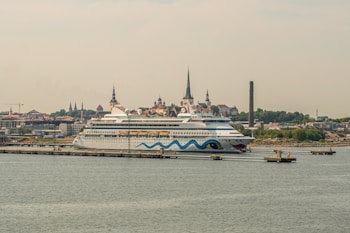 A large cruise ship is docked at a port with a cityscape in the background. The ship features a distinctive design with blue waves and eye motifs on its hull. The city in the background is characterized by several historic buildings with steeples, suggesting a European architectural style. A tall, narrow smokestack is visible, indicating an industrial area nearby. The scene conveys a sense of travel and exploration.