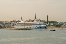 A large cruise ship is docked at a port with a cityscape in the background. The ship features a distinctive design with blue waves and eye motifs on its hull. The city in the background is characterized by several historic buildings with steeples, suggesting a European architectural style. A tall, narrow smokestack is visible, indicating an industrial area nearby. The scene conveys a sense of travel and exploration.