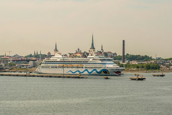 A large cruise ship is docked at a port with a cityscape in the background. The ship features a distinctive design with blue waves and eye motifs on its hull. The city in the background is characterized by several historic buildings with steeples, suggesting a European architectural style. A tall, narrow smokestack is visible, indicating an industrial area nearby. The scene conveys a sense of travel and exploration.
