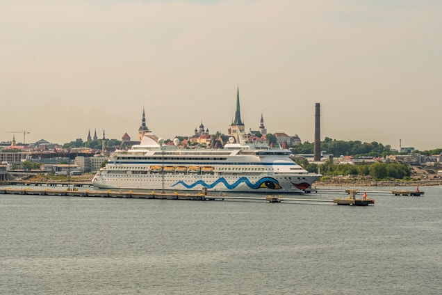 A large cruise ship is docked at a port with a cityscape in the background. The ship features a distinctive design with blue waves and eye motifs on its hull. The city in the background is characterized by several historic buildings with steeples, suggesting a European architectural style. A tall, narrow smokestack is visible, indicating an industrial area nearby. The scene conveys a sense of travel and exploration.