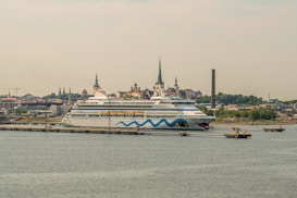 A large cruise ship is docked at a port with a cityscape in the background. The ship features a distinctive design with blue waves and eye motifs on its hull. The city in the background is characterized by several historic buildings with steeples, suggesting a European architectural style. A tall, narrow smokestack is visible, indicating an industrial area nearby. The scene conveys a sense of travel and exploration.