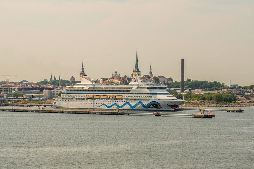 A large cruise ship is docked at a port with a cityscape in the background. The ship features a distinctive design with blue waves and eye motifs on its hull. The city in the background is characterized by several historic buildings with steeples, suggesting a European architectural style. A tall, narrow smokestack is visible, indicating an industrial area nearby. The scene conveys a sense of travel and exploration.