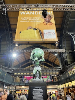A statue of Atlas holding up a globe is positioned in the center of a busy indoor shopping area. Above the statue hangs a large advertisement featuring a smiling person holding a yellow suitcase, with text in German. The setting has an industrial, architectural design with high ceilings and visible steel beams.