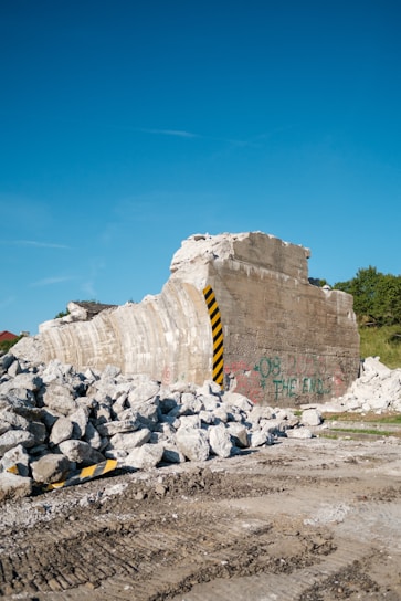 A partially demolished concrete structure with graffiti, surrounded by piles of rubble. The remnants of the structure include exposed rebar and a prominent yellow and black striped warning marker. The sky is clear with a hint of few clouds, and there is greenery in the background.