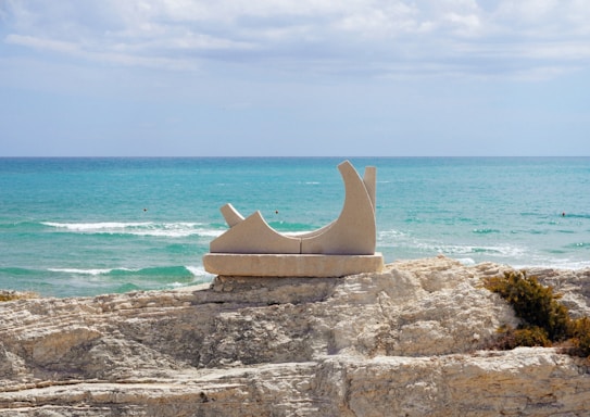 A modern stone sculpture is situated on a rocky coastline, with the vast blue sea and a slightly cloudy sky in the background. The sculpture has an abstract form with smooth curves and angular elements.