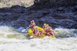 A group of people wearing life jackets and helmets are white water rafting on a yellow raft through rapids. The scene is surrounded by rocky terrain and churning water.