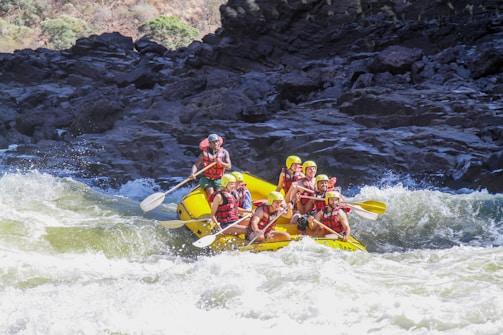 A group of people wearing life jackets and helmets are white water rafting on a yellow raft through rapids. The scene is surrounded by rocky terrain and churning water.
