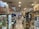 A bustling indoor market aisle filled with various products and merchandise on display. Shelves and display cases hold a variety of items, including gaming accessories, toys, and collectibles. People walk through the aisle, browsing the merchandise, with some customers stopping to examine products closely.