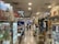 A bustling indoor market aisle filled with various products and merchandise on display. Shelves and display cases hold a variety of items, including gaming accessories, toys, and collectibles. People walk through the aisle, browsing the merchandise, with some customers stopping to examine products closely.