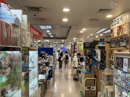 A bustling indoor market aisle filled with various products and merchandise on display. Shelves and display cases hold a variety of items, including gaming accessories, toys, and collectibles. People walk through the aisle, browsing the merchandise, with some customers stopping to examine products closely.