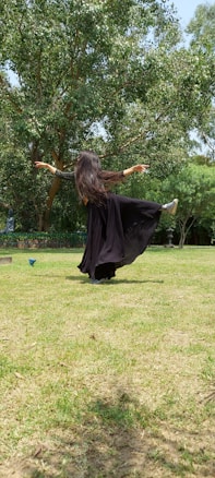 A person wearing a flowing black dress with long hair dances in an open grassy area surrounded by lush green trees. Their arms are stretched outwards, and the dress creates a sense of movement and rhythm.