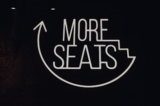 A neon sign with the phrase 'MORE SEATS' is displayed on a dark background. The words are engaging and the design includes an upward arrow around the text, suggesting direction or movement towards a seating area.