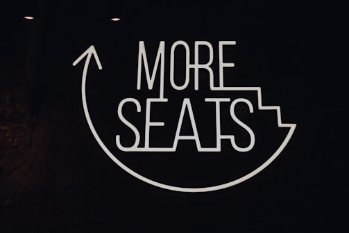 A neon sign with the phrase 'MORE SEATS' is displayed on a dark background. The words are engaging and the design includes an upward arrow around the text, suggesting direction or movement towards a seating area.