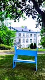 A serene garden setting featuring a blue bench prominently placed in the foreground. The backdrop consists of a grand, elegant building with multiple windows and a light-colored facade. Lush greenery, including trees and bushes, surrounds the area, contributing to a peaceful and inviting atmosphere.